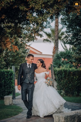 A wedding photo shows a bride and groom walking in the gardens of Torre Giulia, a reception venue in Cerignola, Italy. The couple is strolling through the grounds.